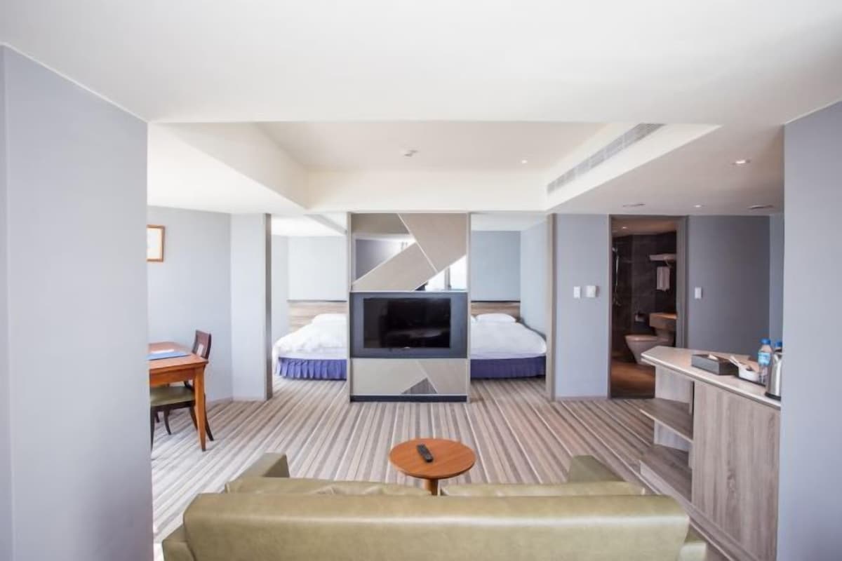 A modern hotel room with a sofa, a TV, a bed, a dining area, and a bathroom.