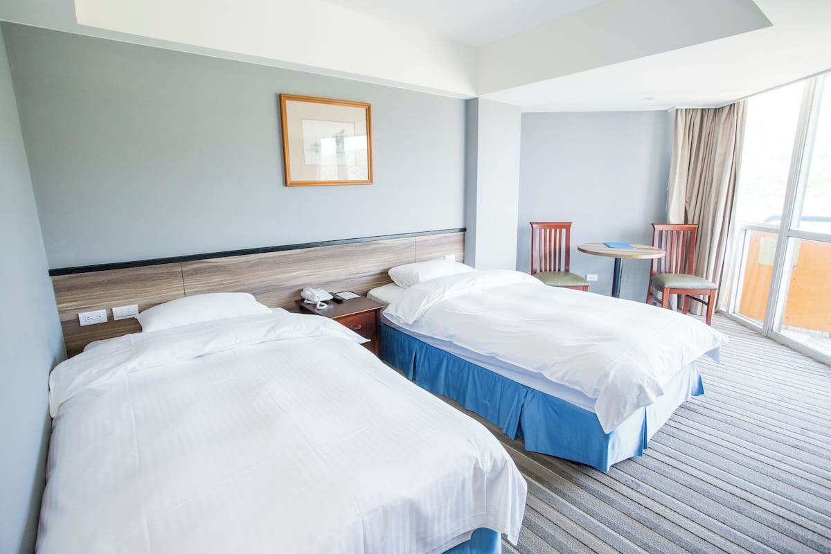 A hotel room with two beds, a small table, a chair, and a window with curtains.
