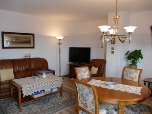 Uma sala de estar com um sofá de couro marrom, uma mesa de centro com toalha de renda, uma mesa de jantar com cadeiras florais, um candelabro, uma televisão e um quadro na parede.