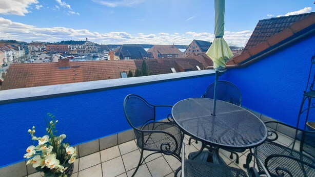 Una terrazza sul tetto con un muro blu, un tavolo di metallo rotondo, due sedie, un ombrellone verde e una pianta in vaso.