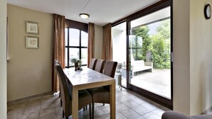 A dining area with a table and chairs, a large window with curtains, and a view of greenery outside. - Holiday house "White Shell" - large garden - parking lot directly at the house  (Brouwershaven)