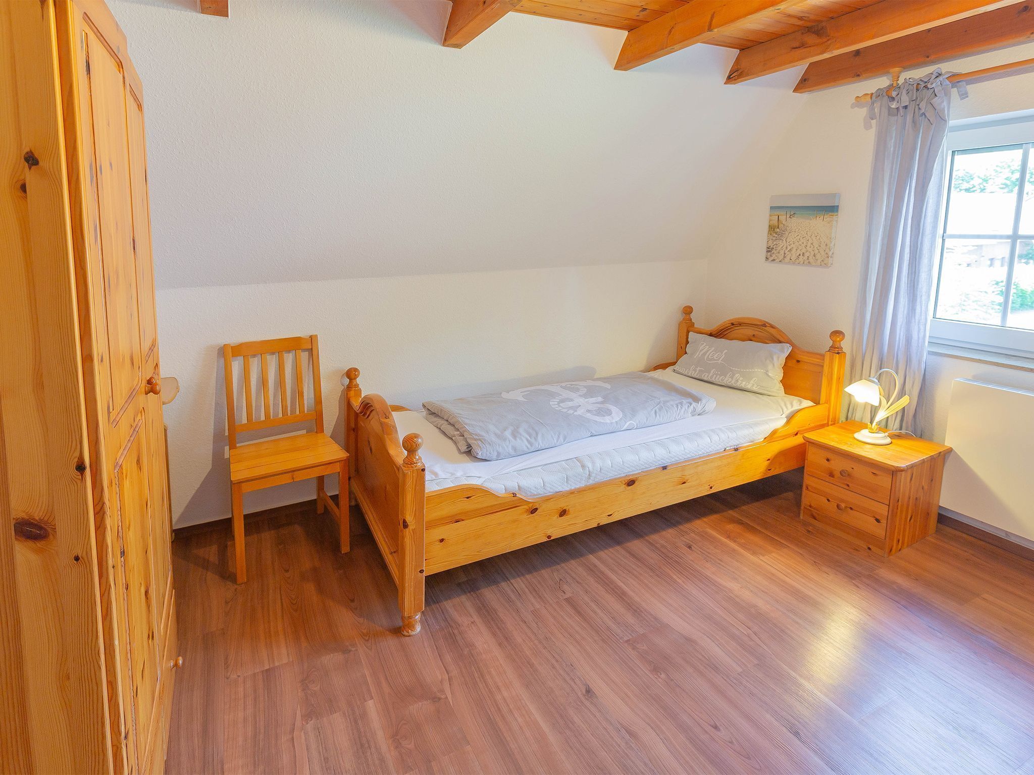 Un lit en bois, avec une table de chevet et une chaise, dans une pièce dont le sol est recouvert de planches de bois et qui possède un plafond en pente.