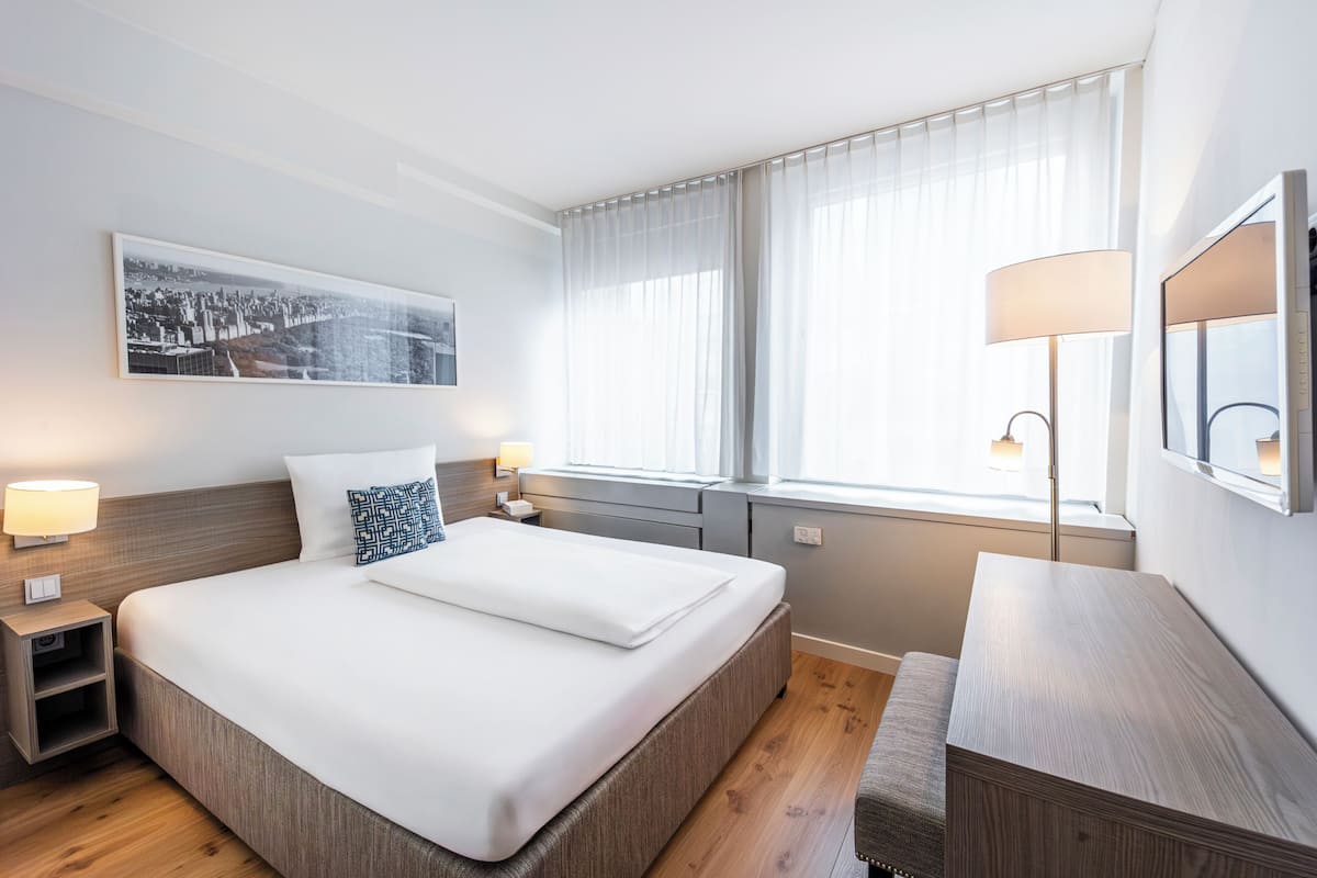 Ein Hotelzimmer mit einem großen Bett, Nachttischen, einem Schreibtisch, einem Sessel, einem Fenster mit Vorhängen, einem an der Wand befestigten Fernseher und einem gerahmten Bild.