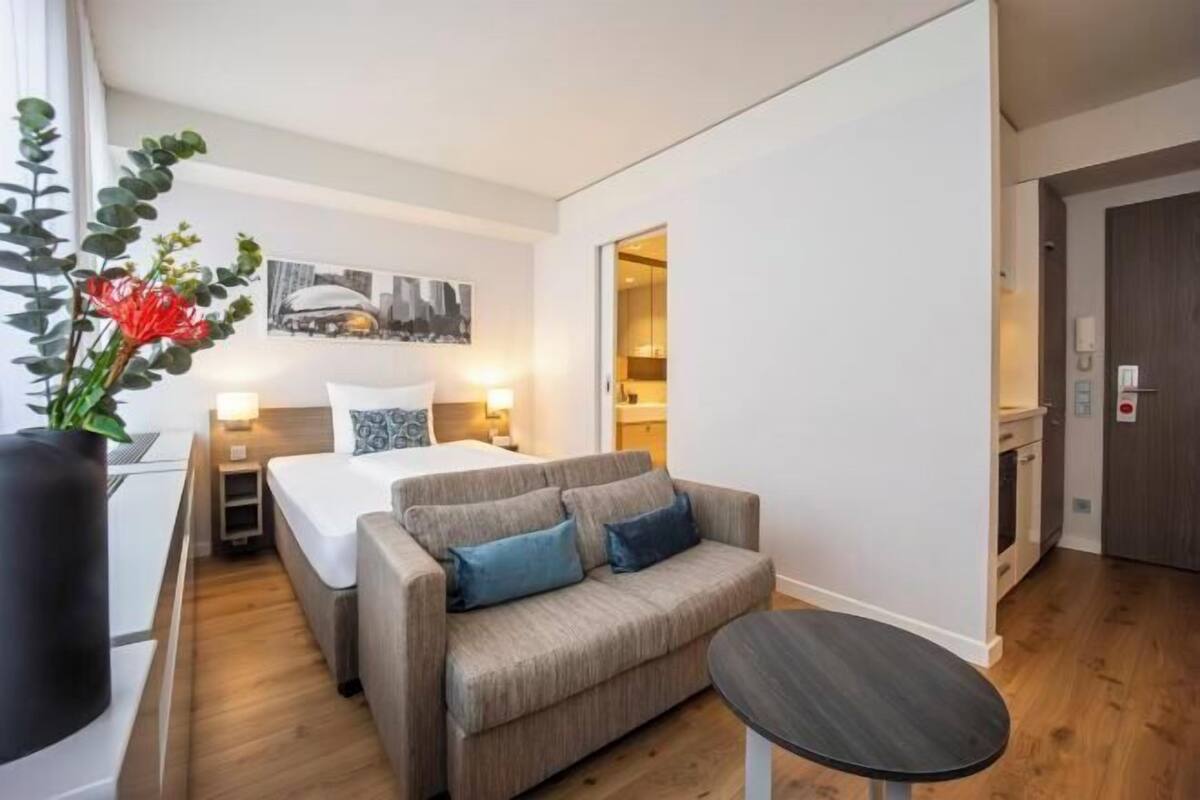 Ein modernes Hotelzimmer mit Bett, einer Couch, einem kleinen Tisch, einer Topfpflanze und einem Stadtbild an der Wand.