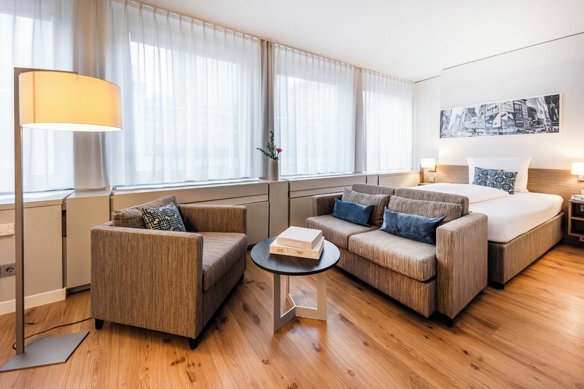 Ein modernes Hotelzimmer mit einer Couch, einem kleinen Tisch, einem Bett und einem großen Fenster mit transparenten Vorhängen.