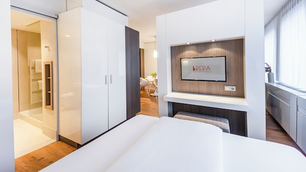 A modern hotel room with a large bed, built-in wardrobe, a TV mounted on the wall, and a dining area visible through an open door.