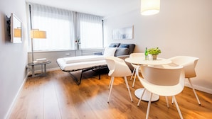 A modern hotel room with a sofa bed, a dining table with chairs, a small side table, and a potted plant.