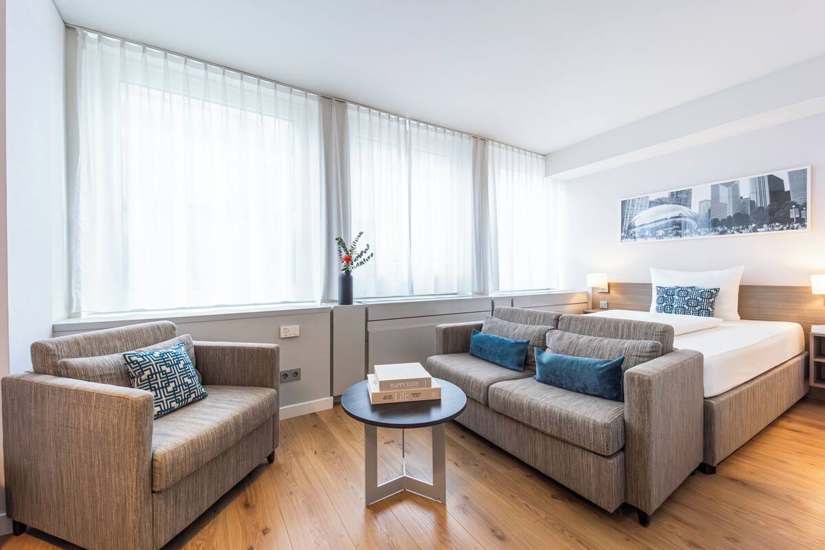Ein modernes Hotelzimmer mit zwei Sofas, einem kleinen runden Tisch, einem Bett mit Stadtbild-Wandkunst und großen Fenstern mit durchsichtigen Vorhängen.