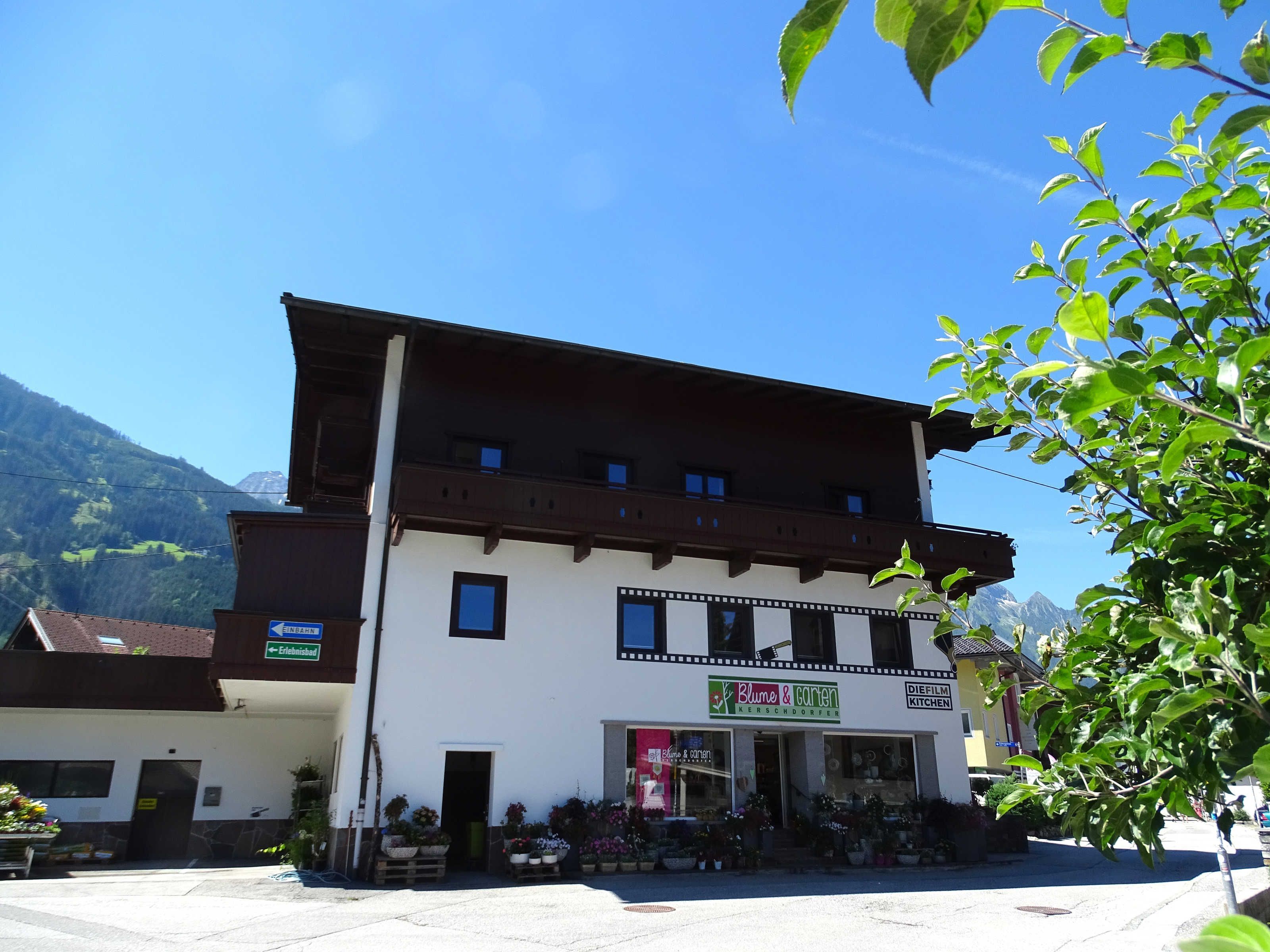 Ein zweistöckiges Gebäude mit weißer Fassade, einem Balkon und einem Schild für 'Blume & Garten', das einen Blumenladen kennzeichnet.