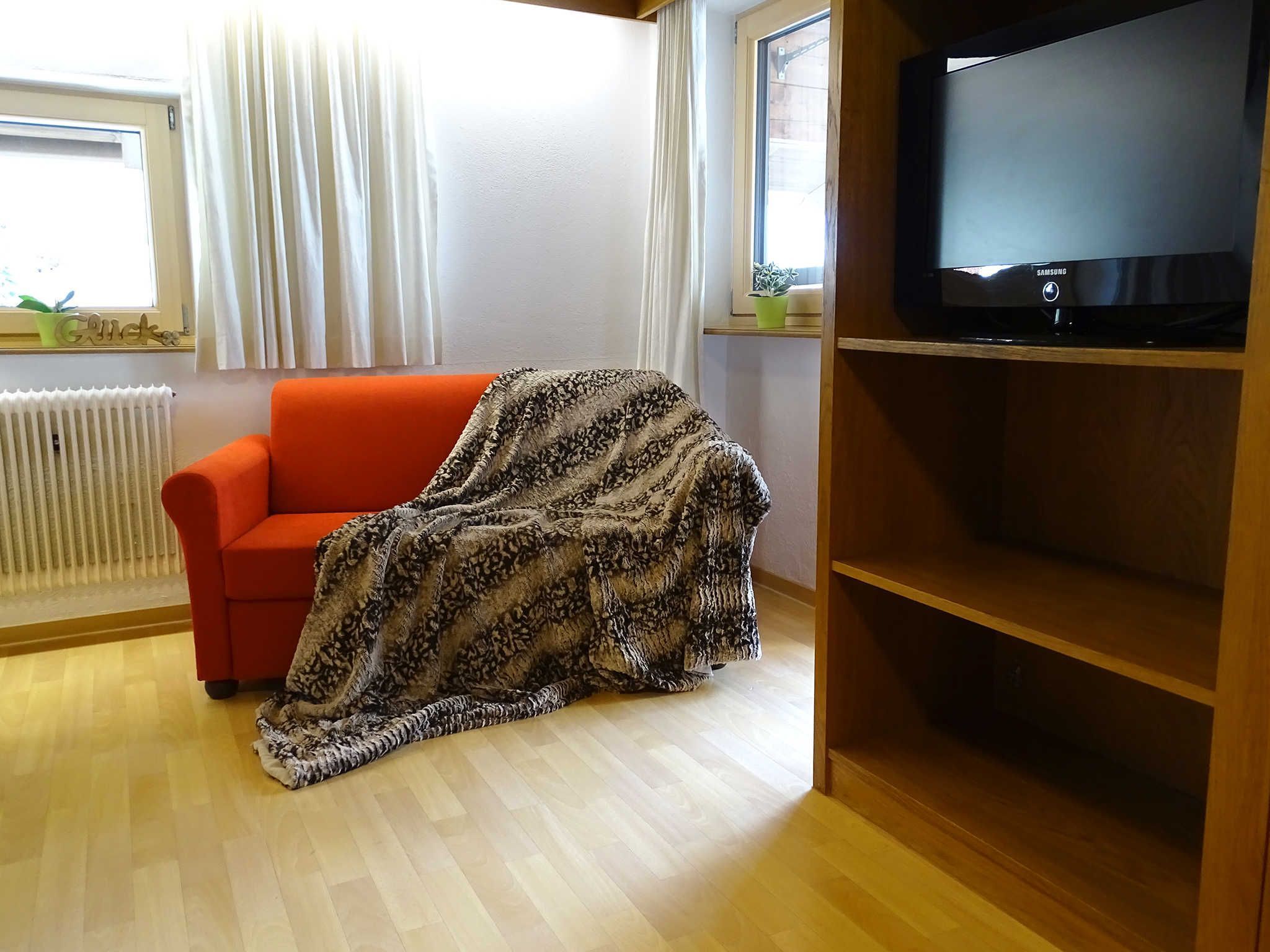 Un salon avec un canapé rouge, une couverture, un téléviseur posé sur un meuble en bois et une fenêtre avec des rideaux.