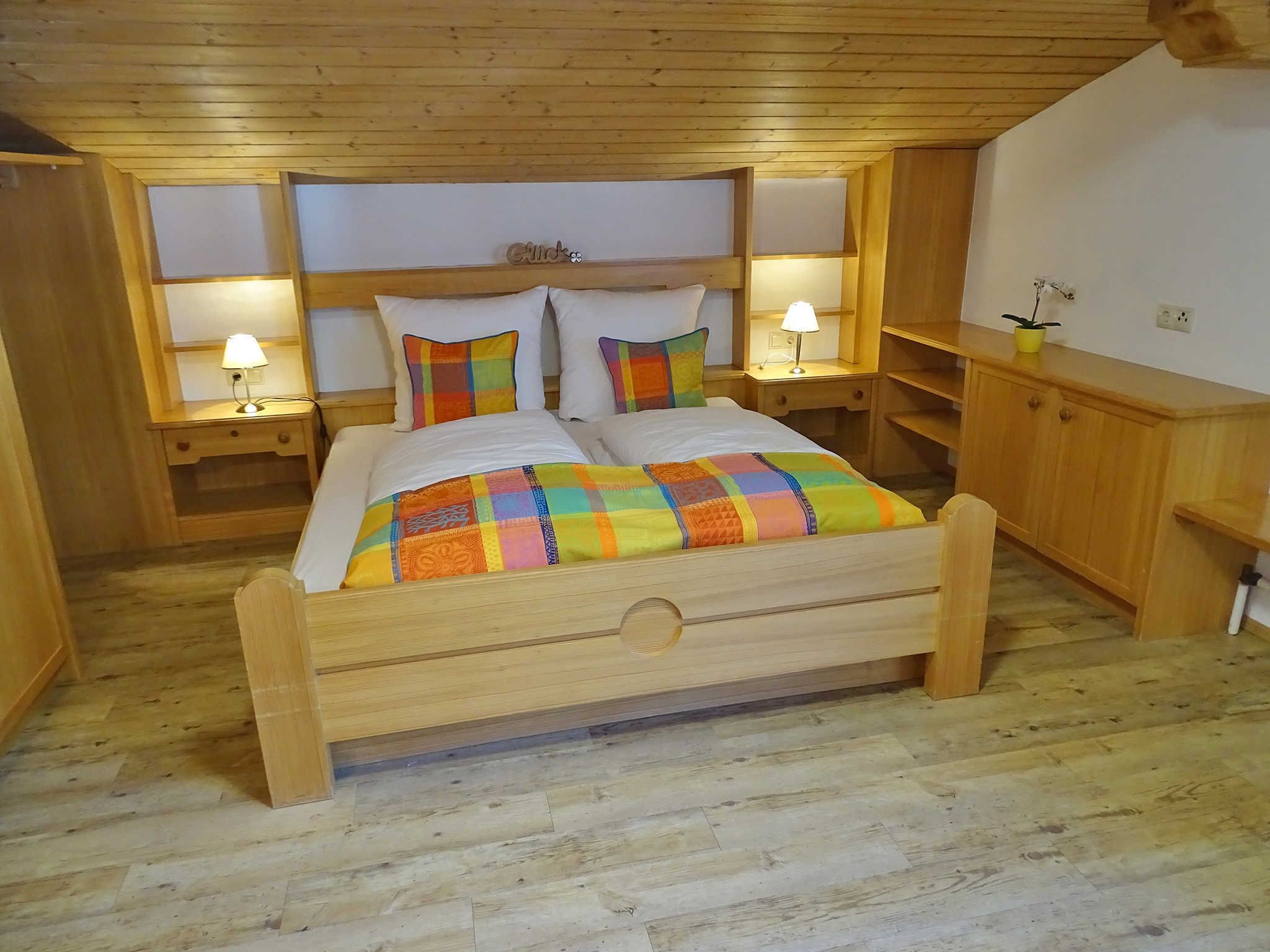 Een houten bed met een kleurrijke quilt, twee bedlampen en een hoofdeinde met een naamplaatje.