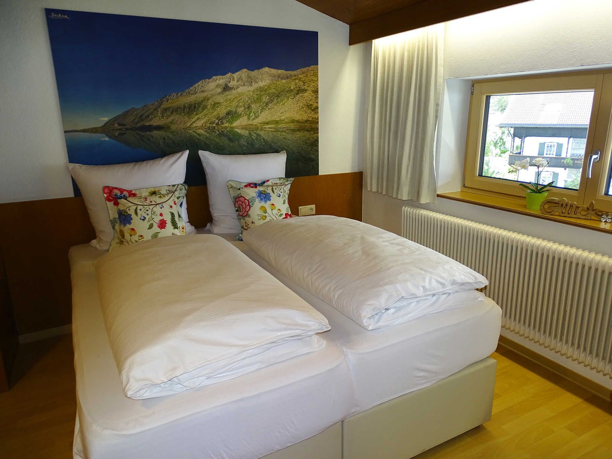 A double bed with white linens, a mountain landscape painting, a window with curtains, and a radiator.