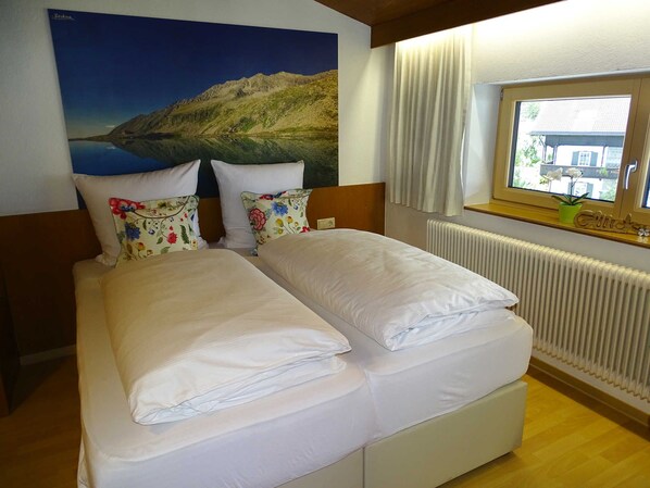 A double bed with white linens, a mountain landscape painting, a window with curtains, and a radiator.