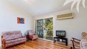 A living room with a wooden floor, two patterned sofas, a television on a stand, and a large sliding glass door leading to a balcony with trees. - Lakeside Retreat @ Smiths Lake (Smiths Lake)