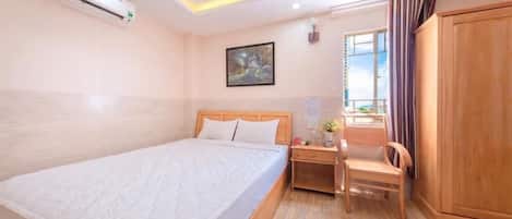 A hotel room with a bed, a desk, a chair, an air conditioner, a painting on the wall, and a wardrobe.