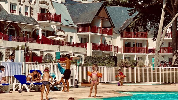 Una piscina con gente en el agua y en el borde. Hay sillas de descanso y sombrillas.