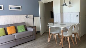 Una sala de estar moderna con un sofá gris, una mesa de comedor con sillas blancas y una zona de cocina con muebles blancos.
