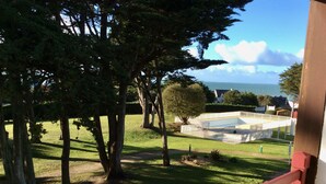 Vista de un área verde con árboles y una estructura blanca, que podría ser una piscina o un patio, con cielo despejado y edificios a lo lejos.