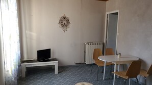 Ein Zimmer mit gefliestem Boden, einem weißen Tisch mit zwei Stühlen, einem kleinen runden Tisch mit einem einzelnen Stuhl, einem schwarzen Fernseher auf einem weißen Schrank und einer Heizung.