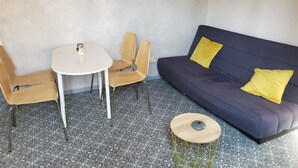 Ein kleiner, moderner Wohnraum mit einem weißen Tisch, zwei Holzsesseln, einem dunkelblauen Sofa mit gelben Kissen und einem runden Holzschemel.