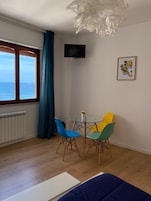 Quarto com piso de madeira, uma mesa redonda de vidro com um vaso, duas cadeiras, uma televisão na parede e uma janela com cortinas.