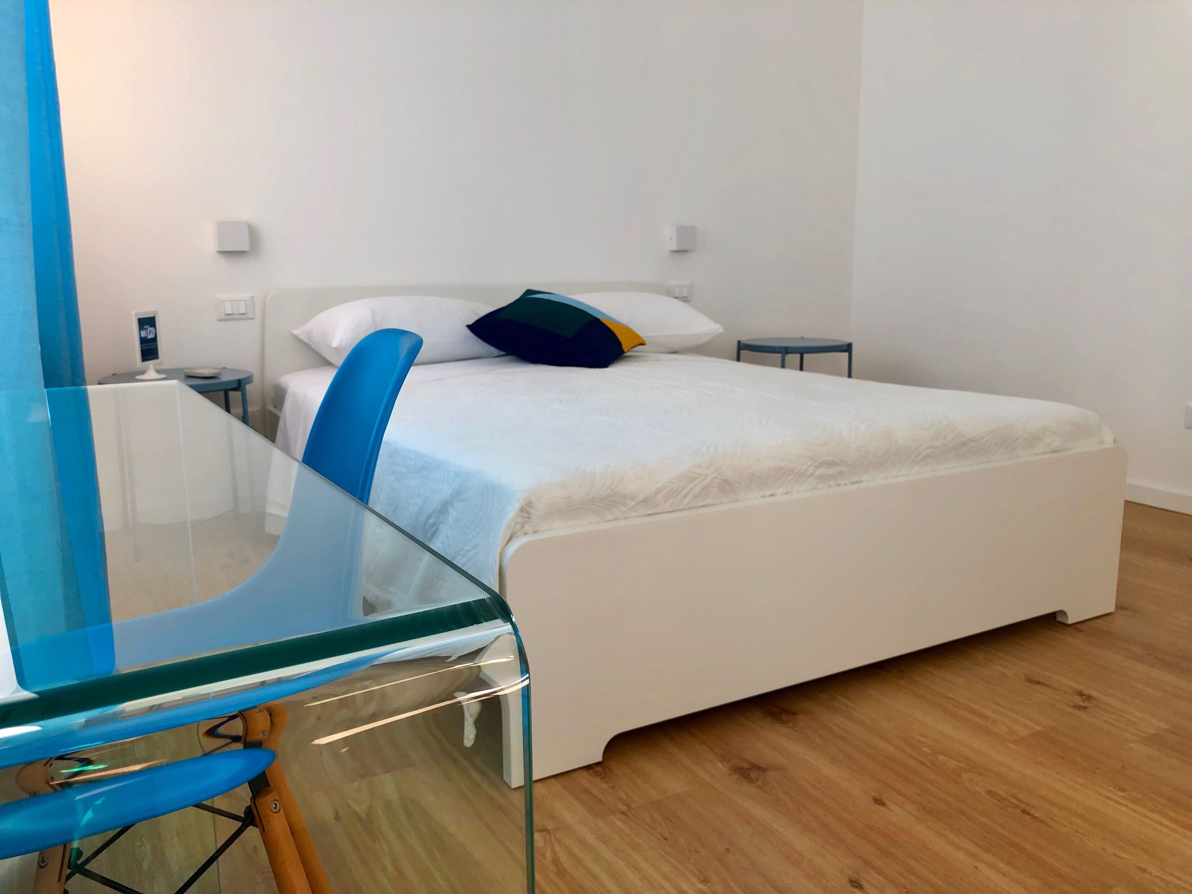 A modern bedroom with a glass desk, a bed with white bedding, and a small bedside table.