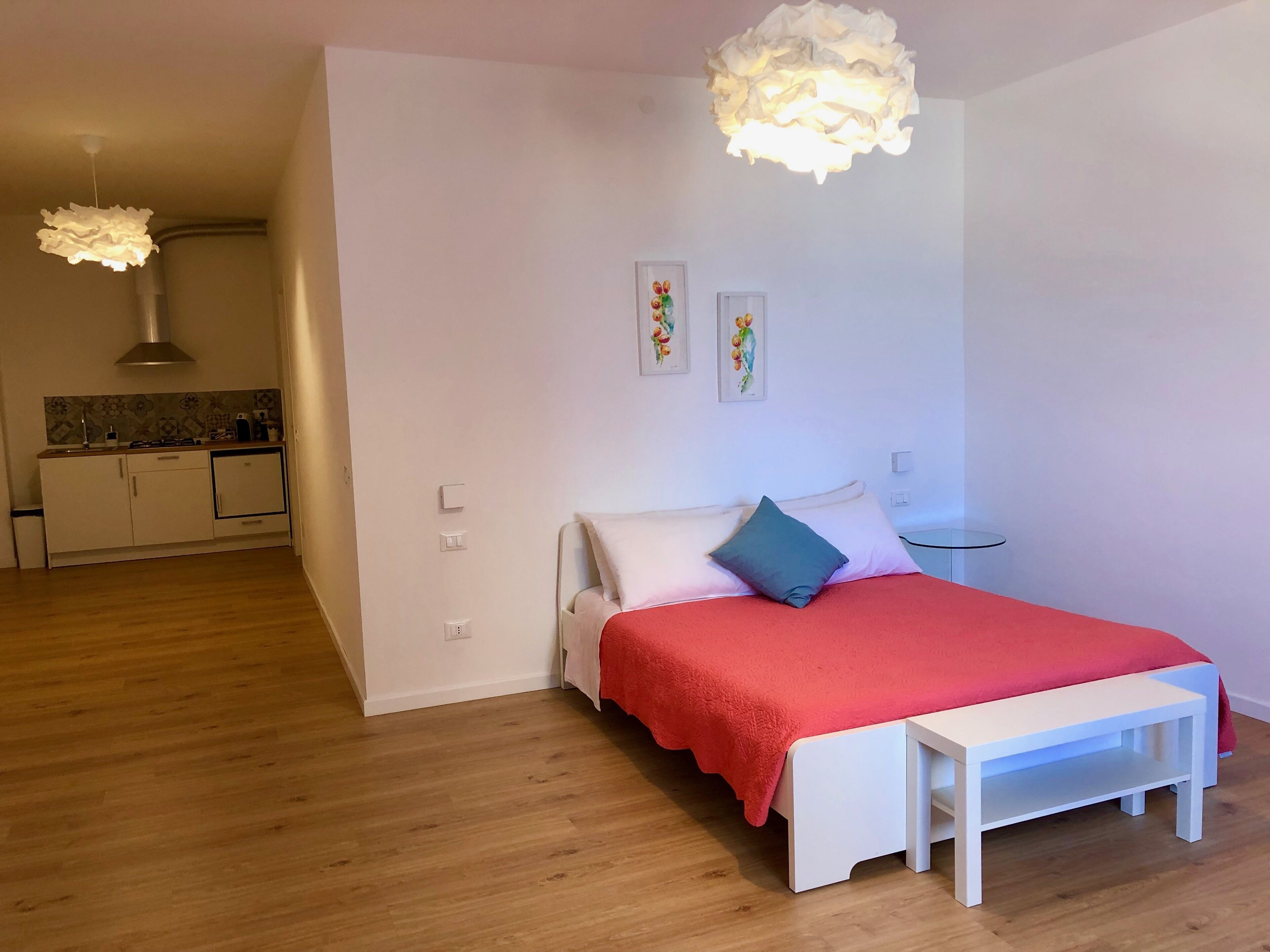 A bedroom with a bed, bedside table, and a red blanket. There is a kitchenette in the background with a stove and cabinets.