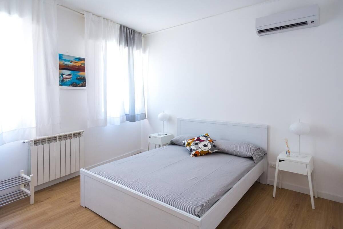 A modern bedroom with a single bed, white walls, a radiator, a window with curtains, a nightstand, a lamp, and an air conditioning unit.