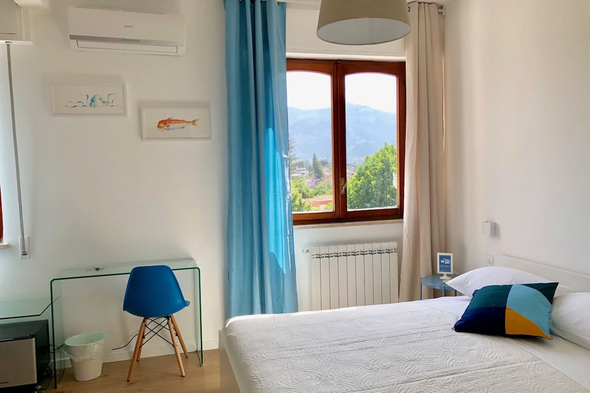 A bedroom with a bed, a desk with a chair, a window with curtains, and a radiator.