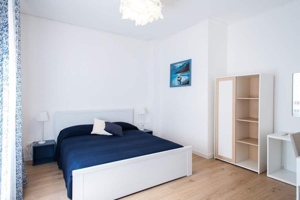 A modern bedroom with a white bed, a nightstand, a wardrobe, and a painting on the wall.