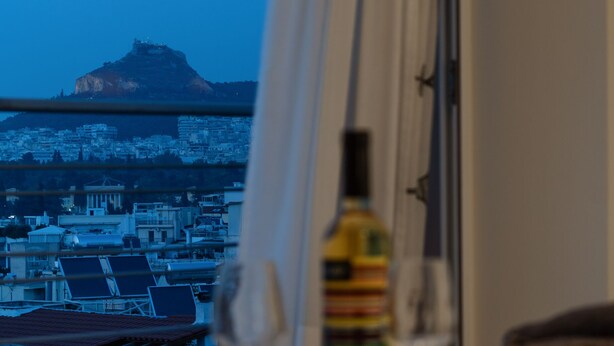 A table with a bottle and a glass, a window with a view of a cityscape and a mountain, and a railing. - Magnificent 76m² homm Penthouse 6th, Acropolis View (Athens)
