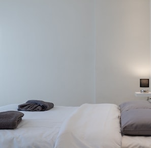 A minimalist bedroom with a bed, pillows, a bedside table with a lamp, and a wall-mounted shelf.
