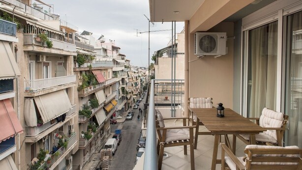 A balcony with a table and chairs, overlooking a street with parked cars and buildings. - Magnificent 101m² homm Apartment, 4th (Athens)