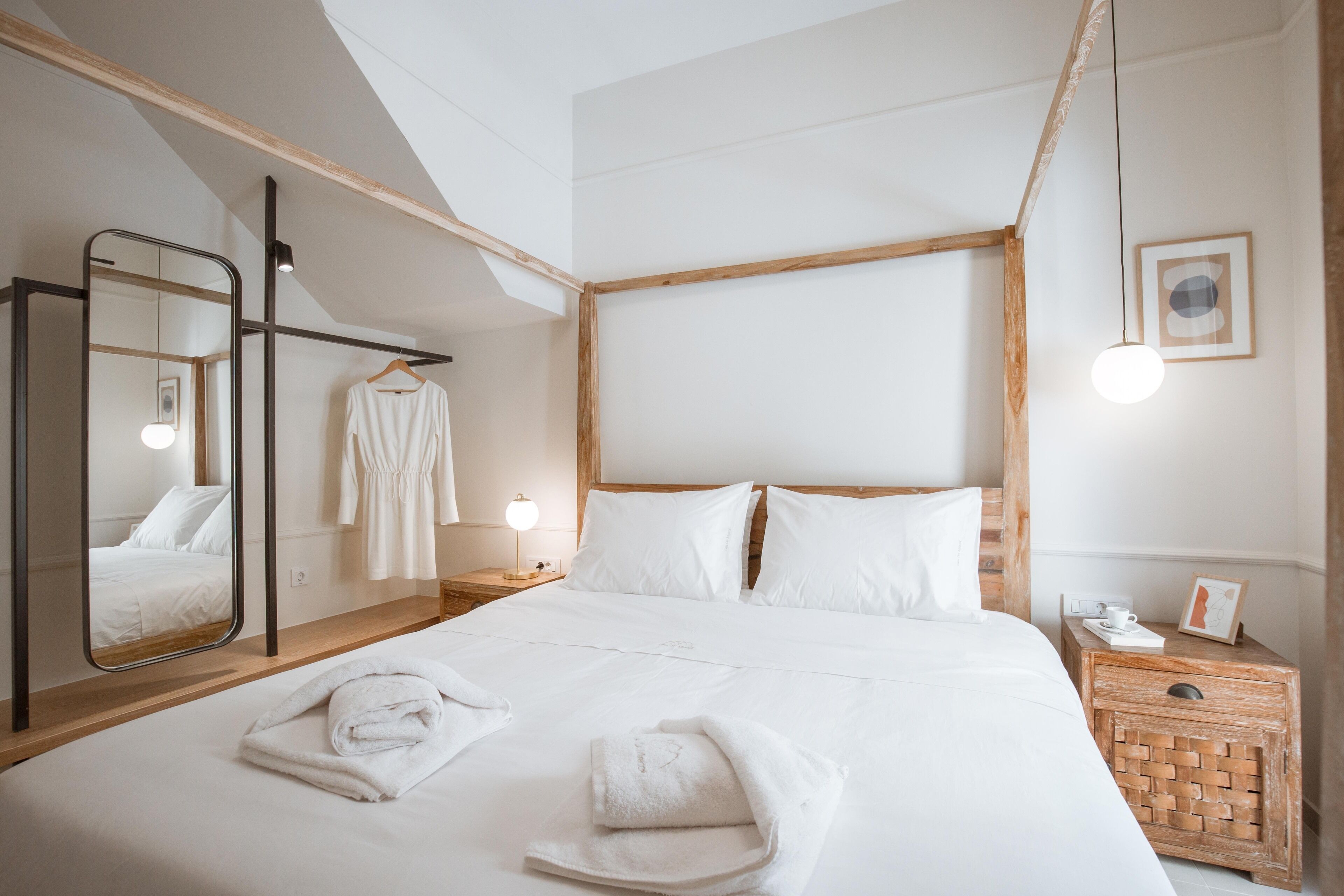 A modern hotel room with a large bed, a wooden headboard, a mirror, a bedside table with a lamp, and a small wooden nightstand.