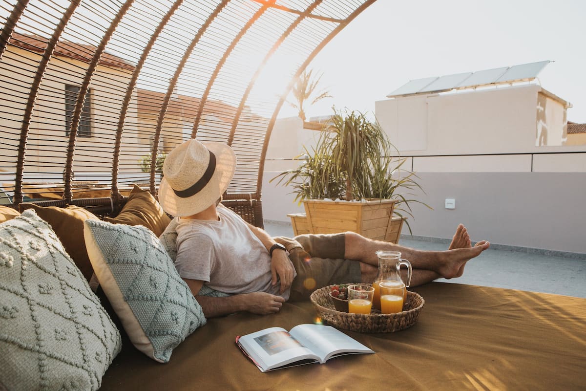 Uma pessoa relaxando em uma área externa com assentos acolchoados, um livro, uma cesta de frutas e um copo de suco de laranja.