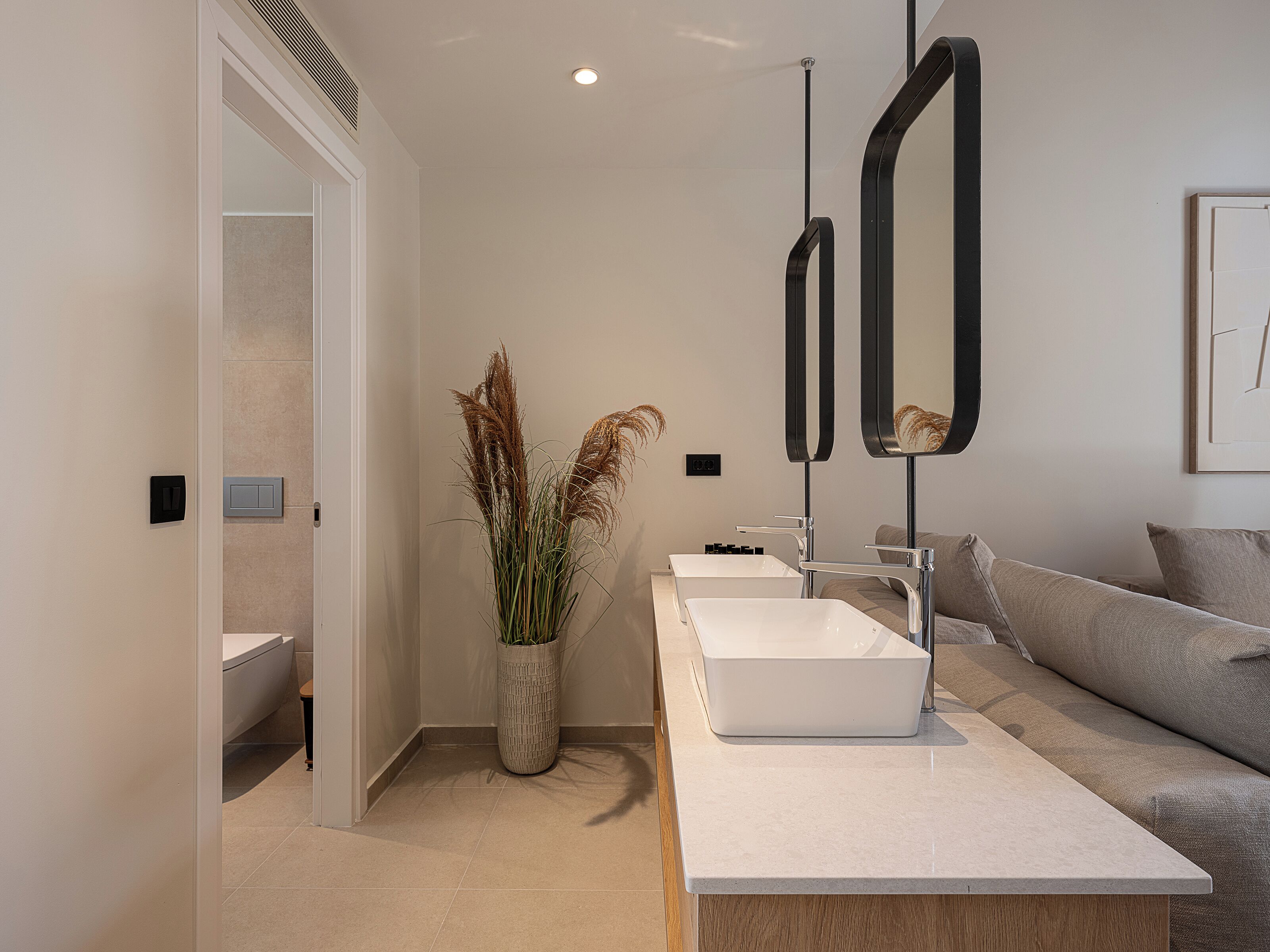 Ein modernes Badezimmer mit einem weißen Waschbecken, zwei Spiegeln und einer Vase mit Trockenpflanzen.