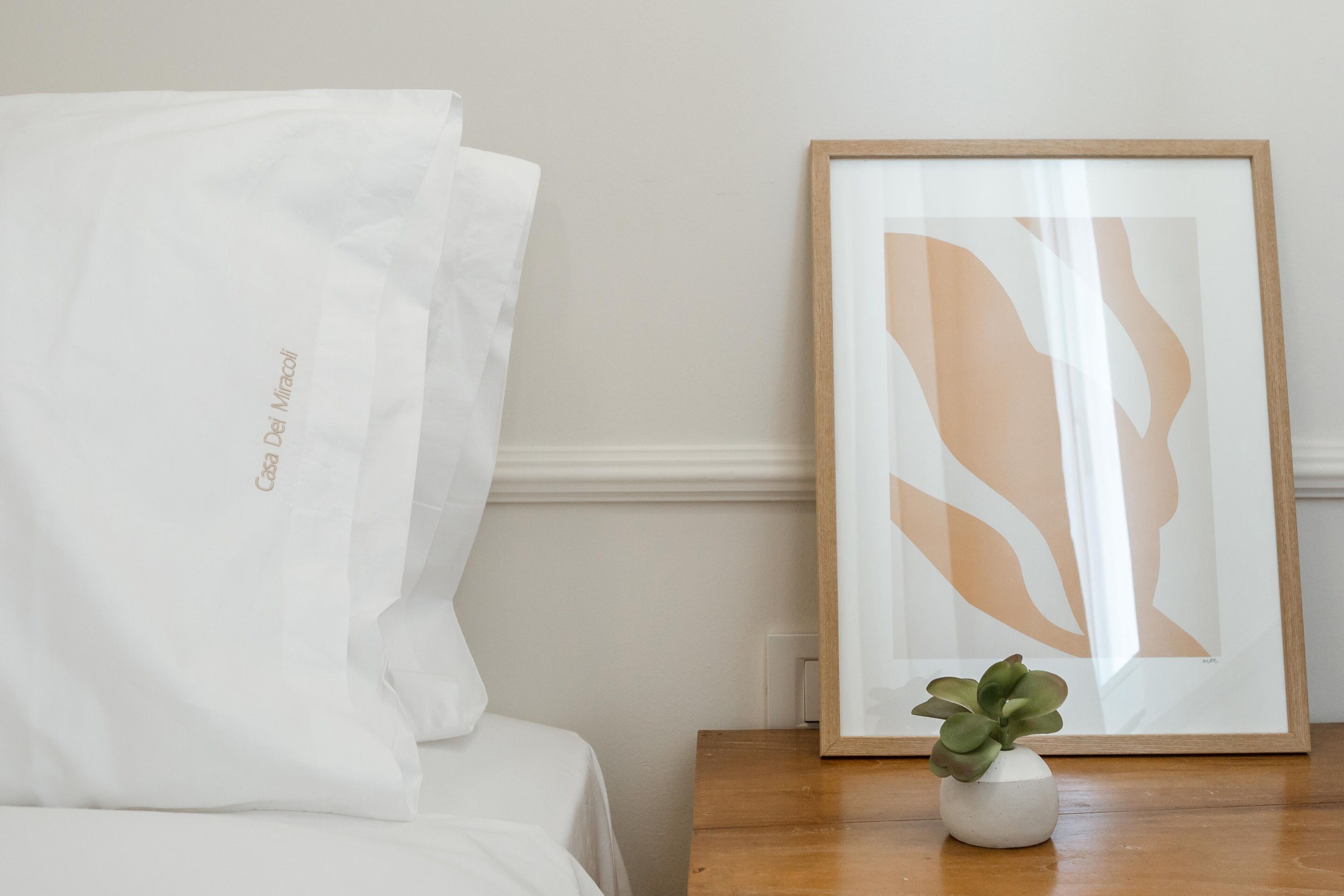 A neatly made bed with a white pillow and a framed abstract art piece on a wooden nightstand.