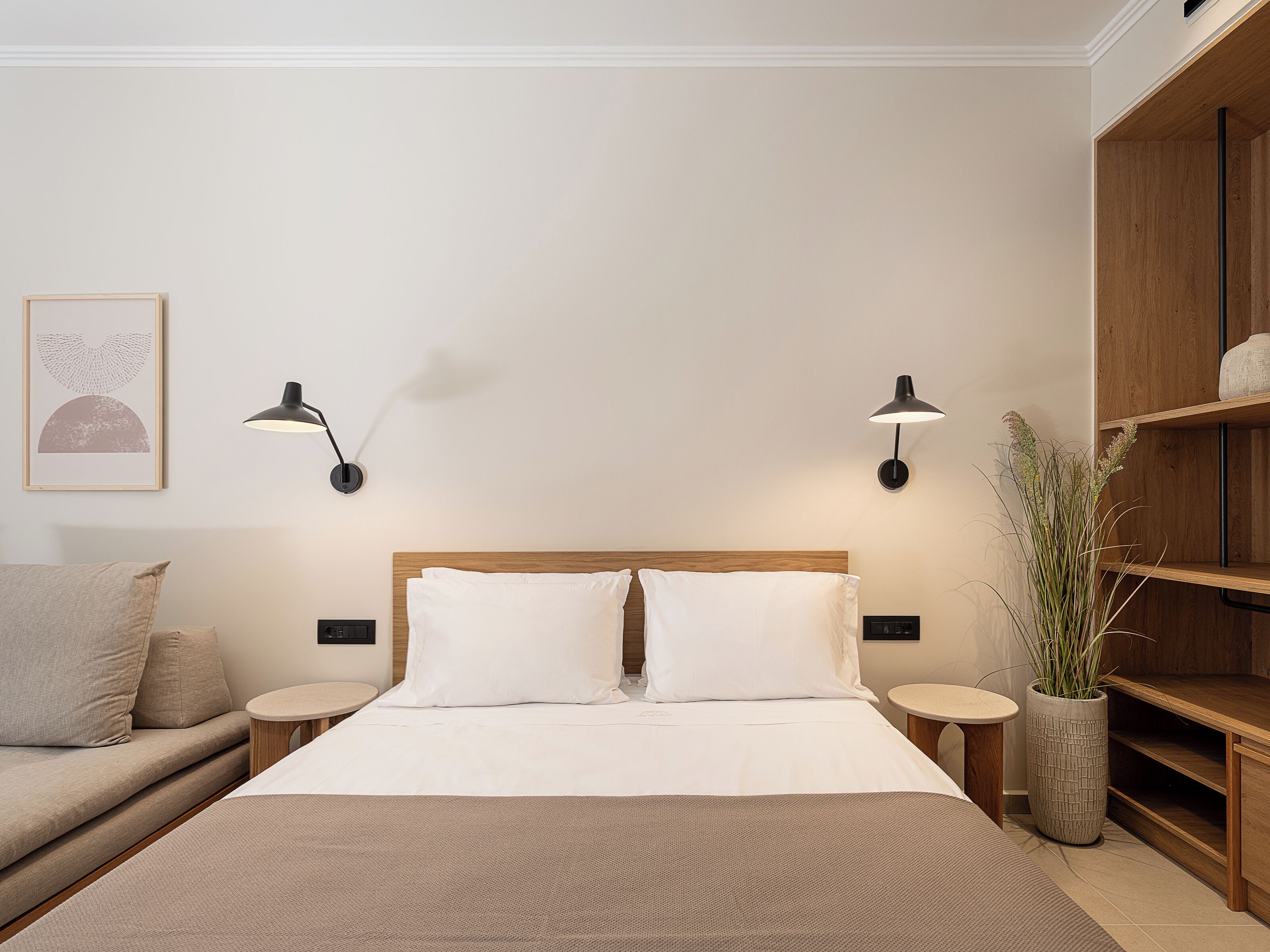 Un dormitorio ordenado con cama, mesitas de noche, una lámpara de pared, una planta en maceta y un estante de madera.