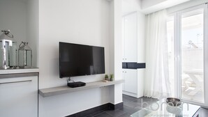 A modern living room with a flat-screen TV mounted on a white wall, a glass coffee table, and a sliding glass door leading to a balcony. - 37.5m² homm Penthouse in Athens with 43m² Terrace (Athens)