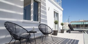 A balcony with two black wicker chairs, a small table with a bottle and glasses, and two potted plants.
