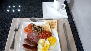 Ein Frühstücksteller mit Toast, Eiern, Tomaten, Pilzen, Würstchen und Bohnen auf einer weißen Untertasse, dazu eine weiße Tasse und Untersetzer.