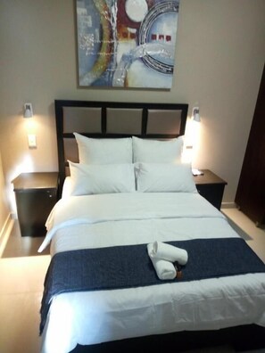 Un lit bien fait, avec du linge de lit blanc et une serviette pliée, une tête de lit ornée d’un tableau, et deux tables de chevet avec des lampes.
