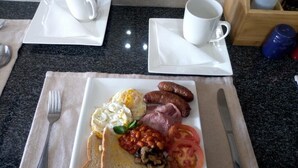 Ein Frühstücksteller mit Eiern, Würstchen, Speck, Tomaten, Bohnen, Pilzen und Toast auf einem Tisch, komplett mit Servietten und Besteck.