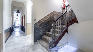 Un couloir au sol de marbre mène à un escalier orné d’une rampe en fer forgé, et un extincteur est fixé au mur.