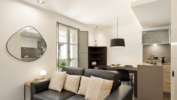 A modern living room with a black leather sofa, a round mirror, and a dining area with a table and chairs. - Modern and comfortable studio in the heart of the city (Barcelona)