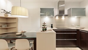 A modern kitchen with a glass dining table, white chairs, dark wooden cabinets, and a hanging light fixture. - Modern and comfortable studio in the heart of the city (Barcelona)