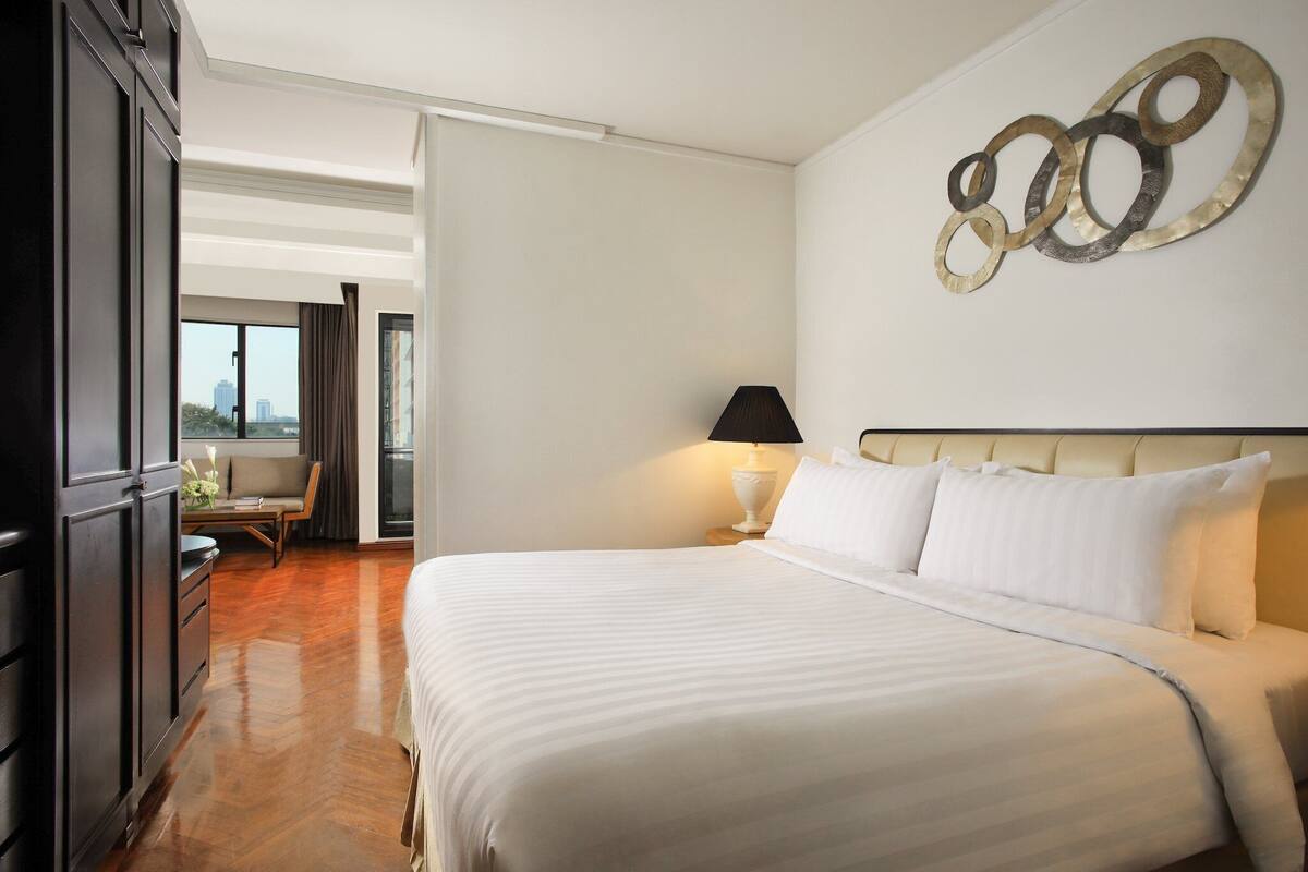A hotel room with a large bed, a nightstand with a lamp, a view of the city through the window, and a decorative wall piece.