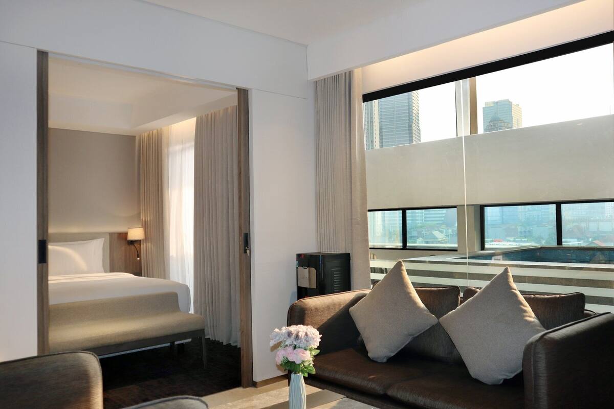 A hotel room with a bed, a sofa, a coffee machine, and a view of the cityscape through large windows.