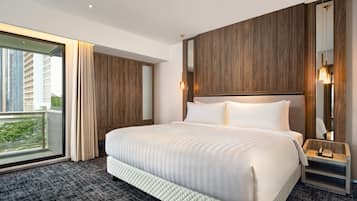 Ett hotellrum med en stor säng, en sänggavel i trä, ett nattduksbord med en lampa och utsikt över byggnader genom ett stort fönster.