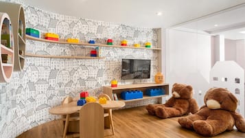 Un cuarto de juegos infantil con una mesa de madera, sillas, un televisor y estantes llenos de bloques de construcción y juguetes.